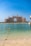 A grand, luxurious hotel with distinctive architecture overlooks the calm blue waters of the sea. The pale sandy beach leads to clear, turquoise water with a line of buoys marking the swimming area. The sky is clear and bright, enhancing the overall elegance and serenity of the scene.