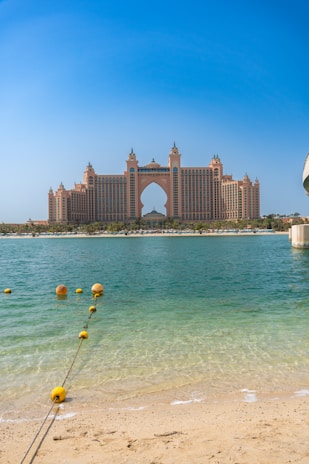 A grand, luxurious hotel with distinctive architecture overlooks the calm blue waters of the sea. The pale sandy beach leads to clear, turquoise water with a line of buoys marking the swimming area. The sky is clear and bright, enhancing the overall elegance and serenity of the scene.