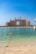 A grand, luxurious hotel with distinctive architecture overlooks the calm blue waters of the sea. The pale sandy beach leads to clear, turquoise water with a line of buoys marking the swimming area. The sky is clear and bright, enhancing the overall elegance and serenity of the scene.