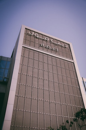 A modern building with a grid-patterned facade, featuring Arabic script signage. The building is captured from a low angle against a clear blue sky.