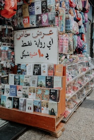 A small street stall displaying an array of books and colorful backpacks. The books are neatly arranged on wooden shelves, featuring a variety of covers and titles. A prominent sign in Arabic hangs above the books, indicating a special offer. The stall is located on a paved street, suggesting a busy market area.