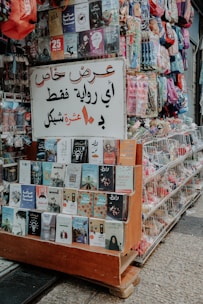 A small street stall displaying an array of books and colorful backpacks. The books are neatly arranged on wooden shelves, featuring a variety of covers and titles. A prominent sign in Arabic hangs above the books, indicating a special offer. The stall is located on a paved street, suggesting a busy market area.
