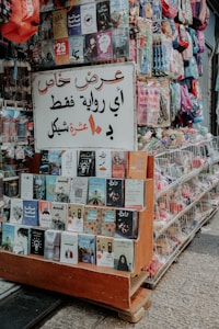 A small street stall displaying an array of books and colorful backpacks. The books are neatly arranged on wooden shelves, featuring a variety of covers and titles. A prominent sign in Arabic hangs above the books, indicating a special offer. The stall is located on a paved street, suggesting a busy market area.
