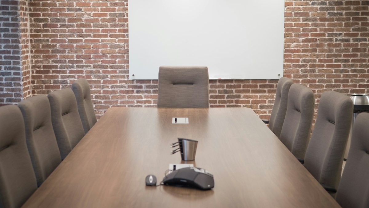 Longue table de réunion vide entourée de fauteuils gris, avec un téléphone de conférence posé au centre et un tableau blanc sur un mur de briques