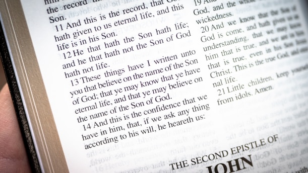An open book displaying a passage from the Bible, specifically from the book of John. The text is clear and readable, with chapter and verse numbers included. The page is predominantly white with black text, and there is a hand holding or touching the book on the left side.