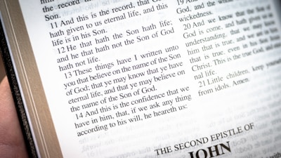 An open book displaying a passage from the Bible, specifically from the book of John. The text is clear and readable, with chapter and verse numbers included. The page is predominantly white with black text, and there is a hand holding or touching the book on the left side.