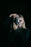 A person with skull makeup is holding a vintage camera, focusing on taking a photograph. The figure is wearing a dark outfit and the background is black, enhancing the mysterious and eerie atmosphere.