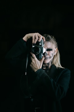 A person with skull makeup is holding a vintage camera, focusing on taking a photograph. The figure is wearing a dark outfit and the background is black, enhancing the mysterious and eerie atmosphere.