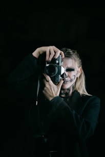 A person with skull makeup is holding a vintage camera, focusing on taking a photograph. The figure is wearing a dark outfit and the background is black, enhancing the mysterious and eerie atmosphere.