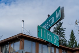 A street sign with two green signs intersecting, indicating Minnesota Avenue and 11th Street. In the background, there is a building with large windows and a chimney. Clear blue sky with scattered clouds and some trees are visible.