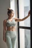 A woman with her hair tied back is dressed in olive green workout attire, standing beside a large window with natural light streaming in. She has one hand resting on the window frame and appears to be looking outside.