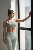 A woman with her hair tied back is dressed in olive green workout attire, standing beside a large window with natural light streaming in. She has one hand resting on the window frame and appears to be looking outside.