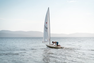 A person in blue shorts is leaning over and adjusting something on a small sailboat with a white sail on a calm sea. The background features distant, hazy mountains under a clear sky.