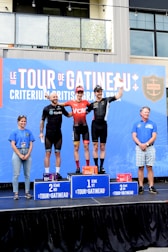 A group of people are standing on a podium, with the top three places marked as first, second, and third. The individuals on the podium are wearing athletic clothing, and the setting seems to be an outdoor event. There is a large banner in the background displaying text related to a tour or race event.
