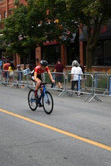 A cyclist wearing a red jersey and helmet rides a blue bicycle along a street lined with metal barricades and spectators. The background features a brick building and trees.