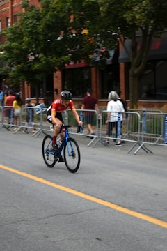 A cyclist wearing a red jersey and helmet rides a blue bicycle along a street lined with metal barricades and spectators. The background features a brick building and trees.