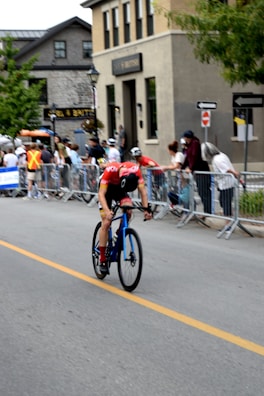 A cyclist in a red and black outfit rides swiftly on a blue road bike along a street lined with onlookers behind barricades. The background includes buildings with multiple floors and some street signs. Spectators, some in casual clothing and others in event staff attire, watch the race.