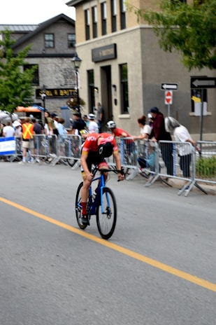 A cyclist in a red and black outfit rides swiftly on a blue road bike along a street lined with onlookers behind barricades. The background includes buildings with multiple floors and some street signs. Spectators, some in casual clothing and others in event staff attire, watch the race.