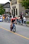 A cyclist in a red and black outfit rides swiftly on a blue road bike along a street lined with onlookers behind barricades. The background includes buildings with multiple floors and some street signs. Spectators, some in casual clothing and others in event staff attire, watch the race.