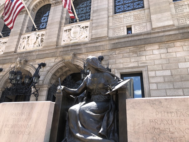 A bronze statue of a seated woman holding a book is prominently positioned in front of an ornate building facade. American flags are displayed above, and inscriptions featuring famous artists and literary figures are carved into the stone walls. Decorative arched windows and wrought-iron lanterns add to the architectural detail.