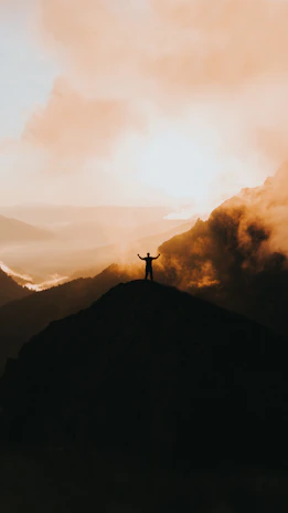 Trekker standing on a mountain peak with arms raised, sunrise in the background.