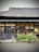 A traditional wooden building with a thatched roof is partially visible. The facade features wooden lattice windows, and a row of yellow flowering plants line the front. The building sits on a gravel surface, adding to the rustic charm. The interior is dimly lit, suggesting a warm interior ambiance.