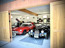 A spacious garage with wooden doors open, revealing two classic cars parked inside. The car on the left is red with a sleek, vintage design. The car on the right is black, also displaying a classic style. The garage interior includes shelves with various items and small windows providing natural light. The floor is concrete, and wooden beams are visible on the ceiling.