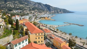 A picturesque coastal town with red-roofed buildings overlooking a turquoise blue sea. The shoreline is lined with palm trees and dotted with small boats in the marina. A mountain range serves as a scenic backdrop, with lush greenery covering the hills.
