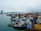 A bustling industrial harbor with numerous tugboats and cargo vessels moored alongside the docks. Cranes and construction equipment are visible in the background, indicating ongoing port activities. The city skyline with modern buildings and a bridge is visible across the water under a cloudy sky.