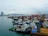 A bustling industrial harbor with numerous tugboats and cargo vessels moored alongside the docks. Cranes and construction equipment are visible in the background, indicating ongoing port activities. The city skyline with modern buildings and a bridge is visible across the water under a cloudy sky.
