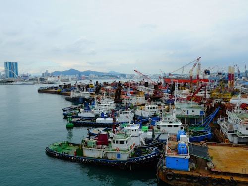 A bustling industrial harbor with numerous tugboats and cargo vessels moored alongside the docks. Cranes and construction equipment are visible in the background, indicating ongoing port activities. The city skyline with modern buildings and a bridge is visible across the water under a cloudy sky.