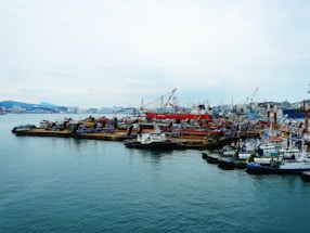 A busy shipyard with numerous ships and cranes, alongside a city skyline in the background. The water is calm, reflecting the industrial activities of the dock. The scene is bustling with maritime vessels, including large and small boats, indicating an active port area.