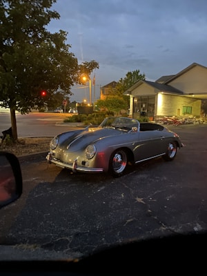 A vintage car is parked outside a building, with evening lights creating a serene atmosphere. The sky appears overcast, and the area is surrounded by a few trees and streetlights. The car's sleek design and chrome details stand out against the darker pavement.