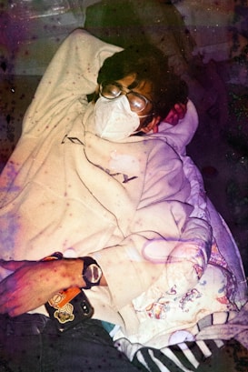 A person is lying down resting, wearing glasses and a face mask. They are dressed in a light-colored hoodie and holding a phone with a visible design on the back. The environment appears cozy, with a patterned blanket and dim lighting contributing to the relaxed atmosphere.