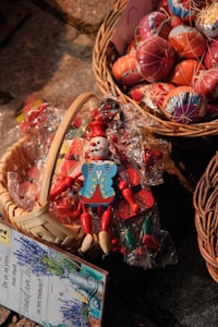 A wicker basket contains a colorful wooden toy with a clown-like figure, alongside packaged items. Nearby, a second basket holds vibrantly decorated Easter eggs. The surface beneath the baskets appears to be cobblestone. A book or pamphlet with lavender imagery and text in Romanian is visible at the edge.