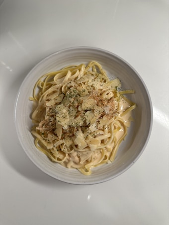 A bowl of creamy pasta is topped with grated cheese and a sprinkle of herbs. The pasta is served in a white bowl, creating a subtle contrast with the pale yellow sauce. The dish has a rustic and comforting appearance, indicating it might be a homemade meal.
