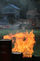 A blaze of fire erupts from barrels next to a body of water. In the background, there are two rustic buildings, one with a visible wooden facade and window shutters, surrounded by trees. The scene is dramatic with smoke rising from the flames.