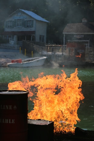 A blaze of fire erupts from barrels next to a body of water. In the background, there are two rustic buildings, one with a visible wooden facade and window shutters, surrounded by trees. The scene is dramatic with smoke rising from the flames.