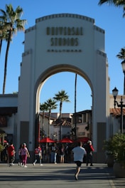 An archway entrance labeled 'Universal Studios Hollywood' with a crowd of people walking beneath it. Palm trees and surrounding buildings with red-tiled roofs are visible in the background under a clear blue sky.
