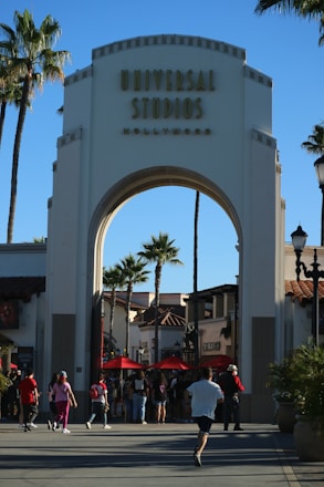 An archway entrance labeled 'Universal Studios Hollywood' with a crowd of people walking beneath it. Palm trees and surrounding buildings with red-tiled roofs are visible in the background under a clear blue sky.