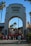 An archway entrance labeled 'Universal Studios Hollywood' with a crowd of people walking beneath it. Palm trees and surrounding buildings with red-tiled roofs are visible in the background under a clear blue sky.