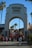 An archway entrance labeled 'Universal Studios Hollywood' with a crowd of people walking beneath it. Palm trees and surrounding buildings with red-tiled roofs are visible in the background under a clear blue sky.