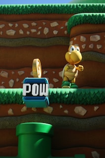 A cartoon-like scene with a yellow turtle character standing on grassy platforms within a layered, earthy background. In the foreground, there is a blue block with the word 'POW' and a green cartoon pipe.