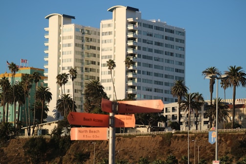 A tall, modern white building is flanked by palm trees against a clear blue sky. In the foreground, there are orange signposts pointing towards various beach locations. Another colorful building with a retro aesthetic is visible to the side.