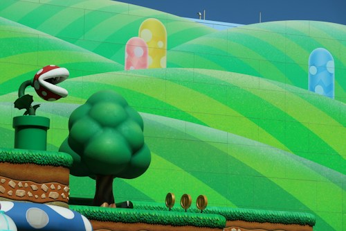 A colorful cartoon landscape featuring a green piranha plant with red and white spots emerging from a pipe. In the foreground, there is a stylized tree and gold coins on a grassy platform. The background includes rolling hills with vibrant green stripes and pastel-colored mushroom-like structures.
