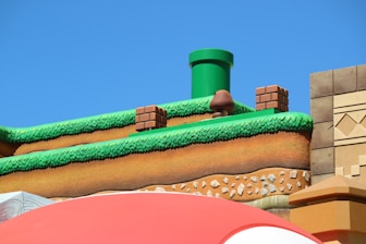 A colorful, stylized scene depicting a platform from a video game setting. The scene features a grass-topped surface with soil layers beneath, a green pipe, brick blocks, and a character resembling a mushroom. The sky is clear and blue, creating a vibrant backdrop.