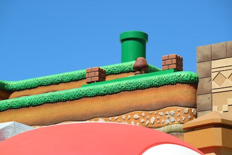 A colorful, stylized scene depicting a platform from a video game setting. The scene features a grass-topped surface with soil layers beneath, a green pipe, brick blocks, and a character resembling a mushroom. The sky is clear and blue, creating a vibrant backdrop.