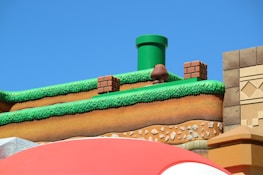 A colorful, stylized scene depicting a platform from a video game setting. The scene features a grass-topped surface with soil layers beneath, a green pipe, brick blocks, and a character resembling a mushroom. The sky is clear and blue, creating a vibrant backdrop.