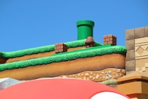 A colorful, stylized scene depicting a platform from a video game setting. The scene features a grass-topped surface with soil layers beneath, a green pipe, brick blocks, and a character resembling a mushroom. The sky is clear and blue, creating a vibrant backdrop.