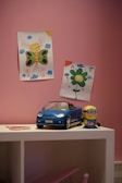 A child's room with pink walls featuring two colorful drawings of a butterfly and a flower on the wall. A small blue toy convertible car and a toy character are placed on a white shelf.